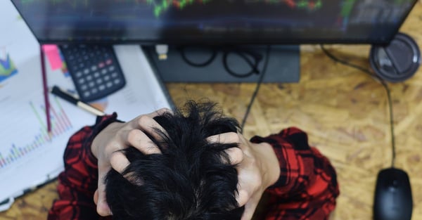 man holding his head between his heads in front of his monitor that displays trading charts